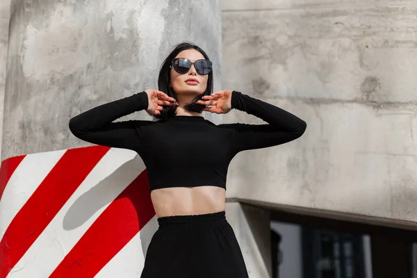 stock image Fashionable beautiful stylish girl model with cool fashion sunglasses in trendy black clothes on the street near the concrete wall with white and red stripes