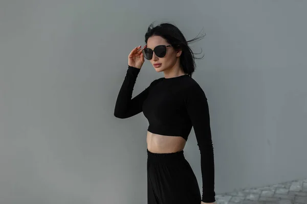 在街上灰蒙蒙的墙上 身穿时髦黑色衣服 戴着时髦黑色太阳镜的漂亮姑娘 — 图库照片