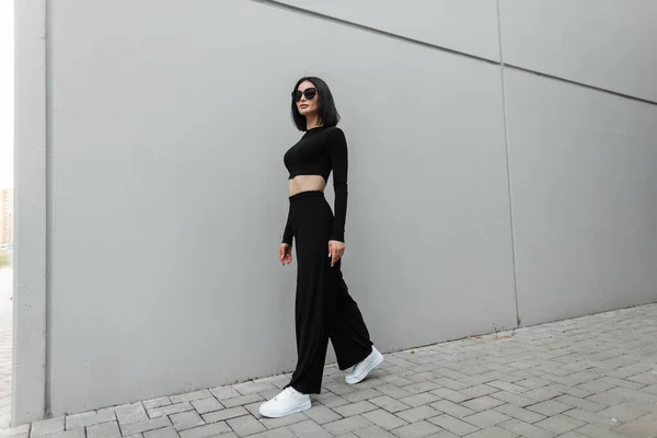 灰色の建物の近くに街を歩く白い靴を持つファッショナブルな黒の通りの服で流行のサングラスとファッション美しい女性モデル — ストック写真