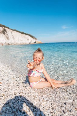 Beautiful happy baby girl in a fashion swimsuit with sunglasses sitting, enjoying and showing the sign of piss on the beach by the blue sea. Summer holidays on island 