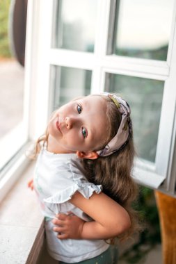 Pencerenin yanındaki kameraya beyaz tişörtlü güzel küçük kız poz veriyor.