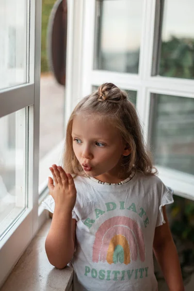 Beautiful little girl in fancy clothes stands by the window and sends an air kiss