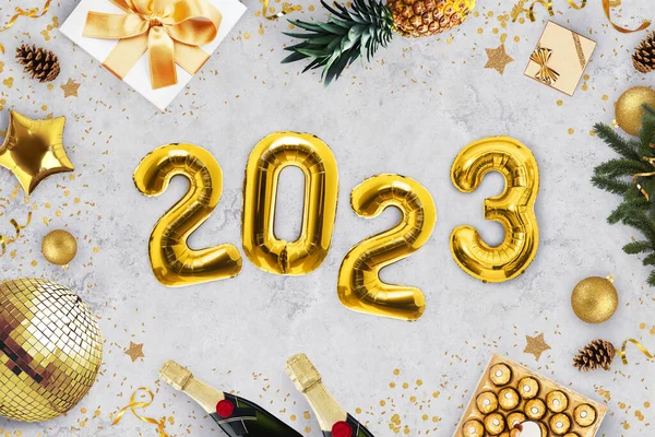 Luxury New Year 2023 party. Golden balloons 2023 lying on gray background with gold mirror ball, candy, champagne, gifts, pineapple, Christmas tree and candy, top view. Holiday celebrations.