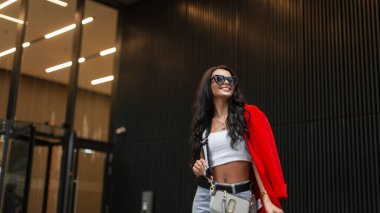 Şık, mutlu, tatlı bir gülümsemesi olan, son moda güneş gözlüklü, kırmızı ceket giyen, beyaz bluzlu ve çantalı güzel bir kız modern bir binanın yanında yürüyor.