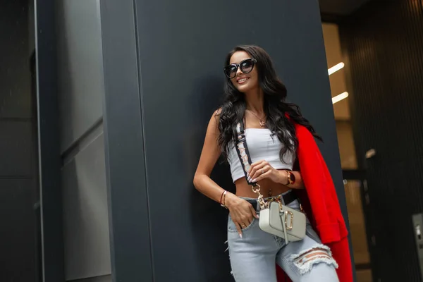 stock image Fashionable beautiful model girl with a smile with curly hair and sunglasses in stylish bright casual clothes with a top, ripped jeans with a belt and a fashion purse stands near a black metal wall