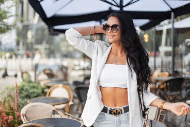 Güzel, mutlu bir iş kadınının portresi. Güneş gözlüklü, şık beyaz elbiseli, güzel bir bayan. Şehirde bir kafe yakınında, bulanık bir arka planda yürüyor.