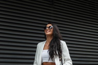 Moda işi güzellik mankeni, modaya uygun güneş gözlüklü, şık beyaz elbiseli, güler yüzlü, şehirde metal bir siyah duvarın yanında yürüyen bir kadın.