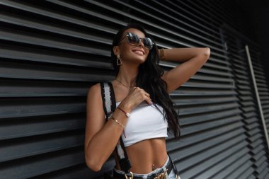 Güzel, güzel bir manken kadın, güzel bir gülümsemesi var, beyaz bir elbisesi var, siyah metal bir duvarın yanında çizgili duruyor. Kentsel kadın tarzı yaz kıyafeti.