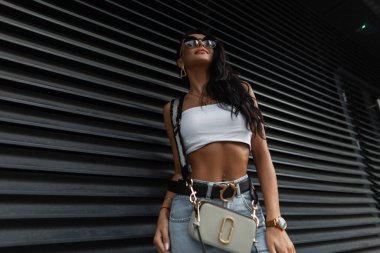 Seksi bronz tenli, şık yaz kıyafetleri içinde, beyaz bluzlu, moda çantalı ve siyah metal bir duvarın yanında güneş gözlüklü şık bir kadın.