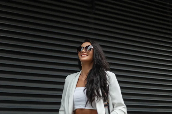 时尚商界美女模特 戴着时髦的太阳镜 身穿时尚的白色休闲装 笑着走在一座金属黑色墙边的城市里 — 图库照片