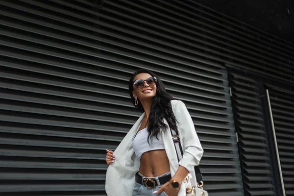 流行の白いカジュアルな服でスタイリッシュなサングラスを持つファッショナブルな美しい幸せな女性のビジネス女性は ブラックメタルの壁の近くに街を歩く バッグ付きのブレザー トップとジーンズと都会の女の子 — ストック写真