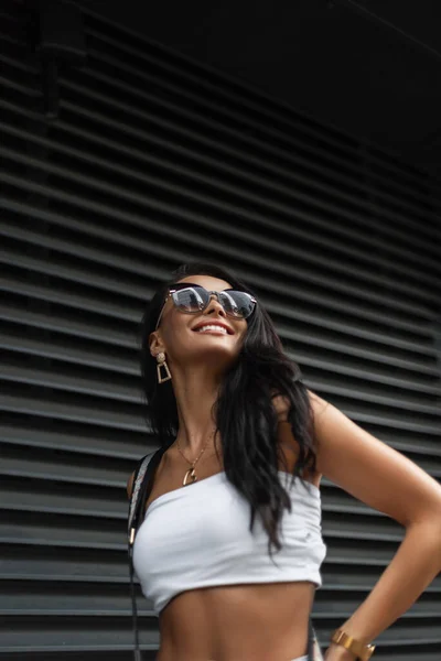 黒い金属の壁の近くの通りを歩くバッグ付きのファッションホワイトトップでサングラスで笑顔で美しいファッショナブルな女の子の幸せな都市の女性の肖像画 — ストック写真