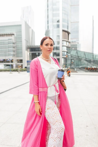 Beautiful stylish plump woman in fashionable bright summer clothes with a pink cape dress and lace pants walks in the city and drinks coffee. Urban fashion style and beauty