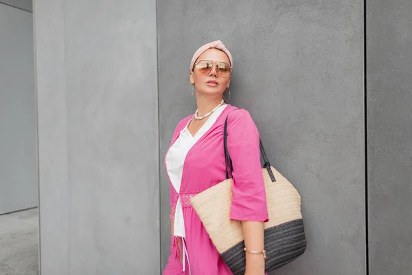 时尚靓丽的女模特 加上时髦的太阳镜 夏天时尚的衣服 粉色的长袍 还有一个大草袋 站在街上灰色的混凝土墙边 — 图库照片