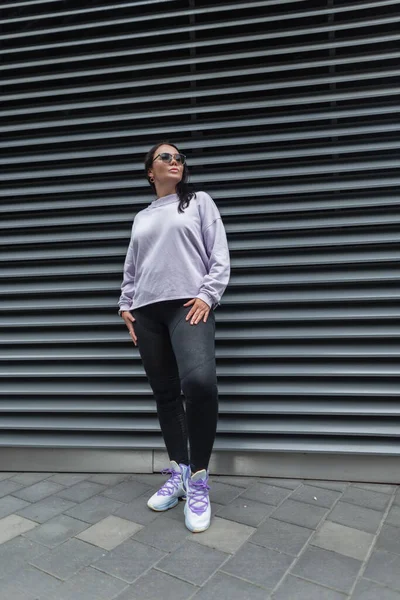 Beautiful fashionable sport plump woman in stylish clothes with a purple oversized pullover with black leggings and trendy sneakers stands near a black metal wall on the street