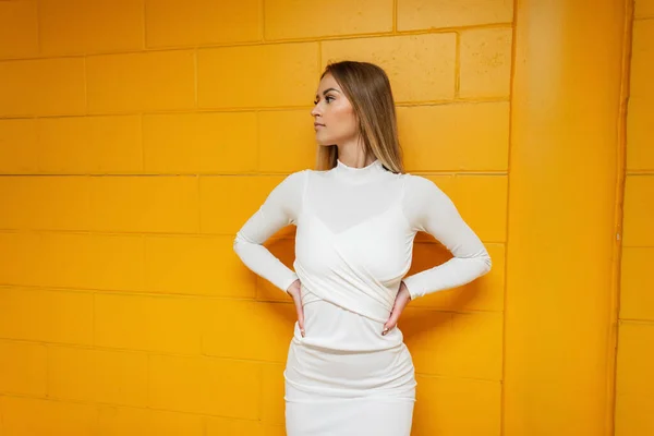 Stylish beautiful girl model in a fashion white dress stands near a yellow brick wall in the parking lot. Urban female style look outfit