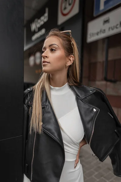 Urban fashion pretty woman in black rock leather jacket and white dress walks on the street