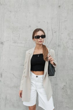 Trendy fashion hipster woman model with sunglasses in stylish casual outfit with blazer, top and shorts with bag stands near a gray wall on the street