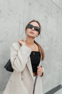 Stylish beautiful young business woman with fashion black sunglasses in casual clothes with a bag on a background of a concrete grunge wall