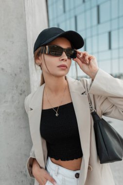 Street portrait of cool trendy young hipster woman with black cap in fashion casual clothes with blazer, top and bag wears sunglasses in the city. Urban girl