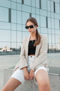 Beautiful woman model with modern sunglasses in fashion street casual clothing with a blazer, top and shorts sits in the city
