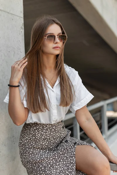 Stylish beautiful business girl in fashion summer clothes with a white shirt and skirt sitting on a metal railing in the city