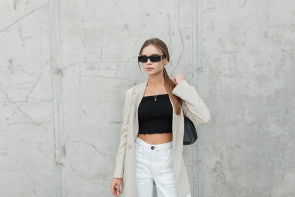 Cool business pretty lady with trendy sunglasses in fashion casual clothes with a blazer, shorts and a top and purse near a gray concrete wall in the city