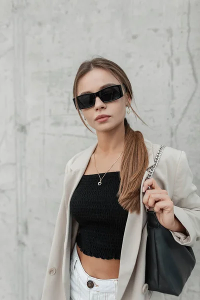 Urban fashion pretty woman with modern stylish black sunglasses in casual outfit with blazer, top and fashionable bag walks on the street near a grey concrete wall
