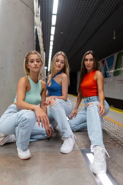 Stylish beautiful young women in fashion clothes with a top and jeans with white sneakers sit and pose in the subway