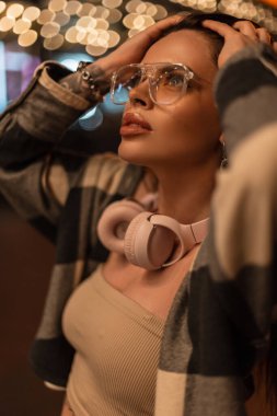 Stylish nighttime female portrait of a beautiful fashionable woman with glasses and rose-colored sunglasses in fashionable clothes looking up walking in the night city and fixing her hair