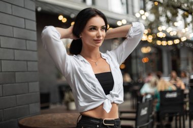 Fashionable young beautiful woman in casual clothes with white shirt stands in the city and fixing hair on a urban background with light bokeh