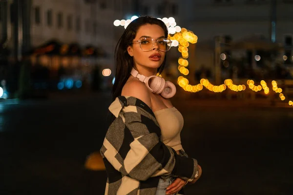 Beautiful sexy fashion woman with big lips with glasses and headphones in fashionable trendy clothes with stylish plaid shirt and top walking in the night city with bokeh lights