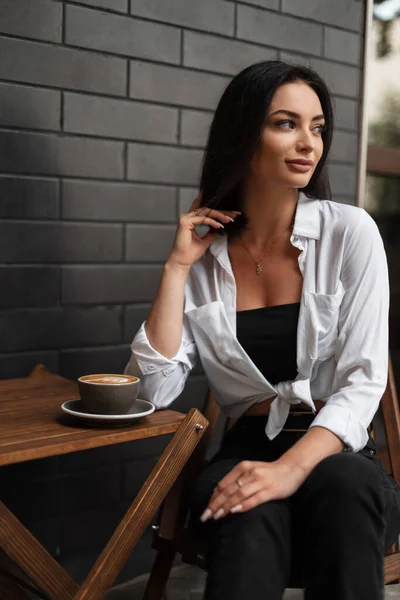 Beautiful woman with white shirt and black jeans sits at table with cup of coffee on the street near a black brick wall.