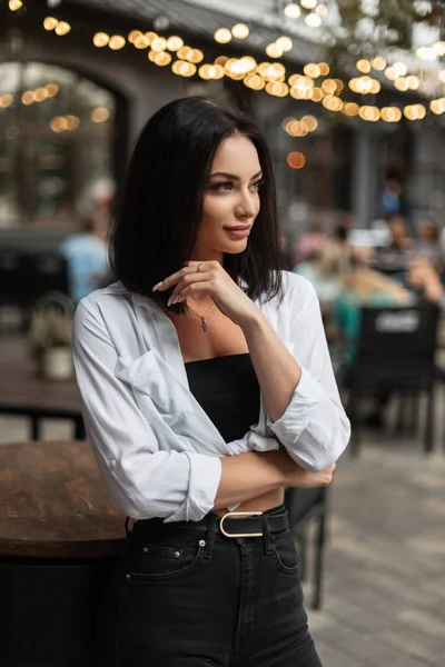 Stylish beautiful successful woman model in fashion clothes with a white shirt stands in the city outside a cafe with bokeh lights