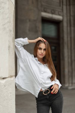 Elegant beautiful young woman model in fashion casual clothes with a white shirt and black jeans stands near a vintage building in the city. Pretty girl