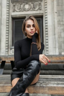 Stylish beautiful young woman model in fashionable black summer clothes with a long sleeve shirt, shorts and high boots sits in the city near a vintage building