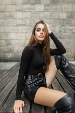 Beautiful fashion girl model with stylish elegant urban black clothes with black long sleeve shirt golf, leather shorts and slender legs with high shoes sits on a wooden bench near brick vintage wall