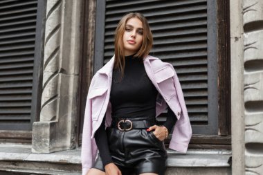 Stylish beautiful young woman model in fashionable black clothes with a pink jacket sits near a vintage building. Trendy elegant pretty girl