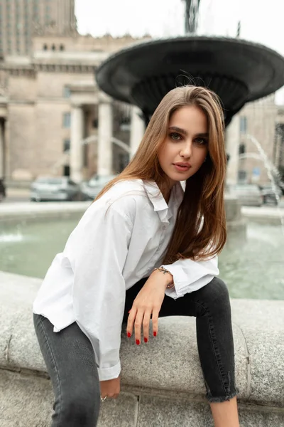 Stylish beautiful vogue model woman in trendy fashion casual clothes with a white shirt and black jeans sits and poses near the fountain in the city