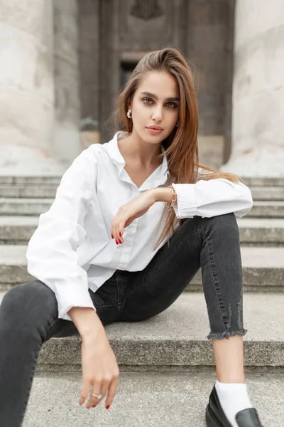 Fashion beautiful woman vogue model in fashionable stylish elegant clothes with white shirt and black jeans sits on the steps in the city and looks into the camera