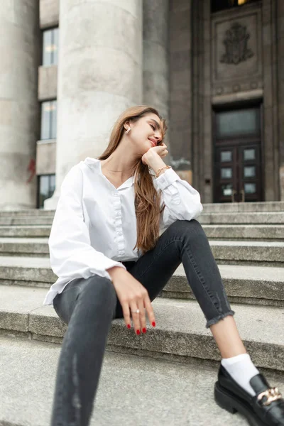 Happy beautiful young woman vogue model in trendy elegant casual clothes with fashion white shirt and black jeans with shoes sits on the steps near the vintage building in the city