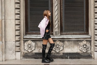 Fashion pretty woman mode in elegant stylish clothes with leather shirt and high boots with pink jacket walks near a vintage wall with window