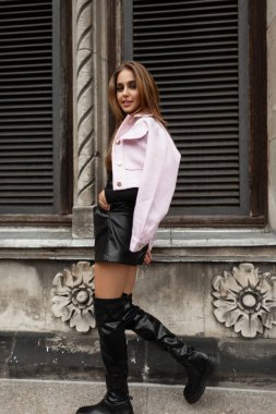 Cool trendy glamorous beautiful woman in stylish elegant clothes with leather shorts with black fashion high boots and a pink jacket walks near a vintage building in a european city
