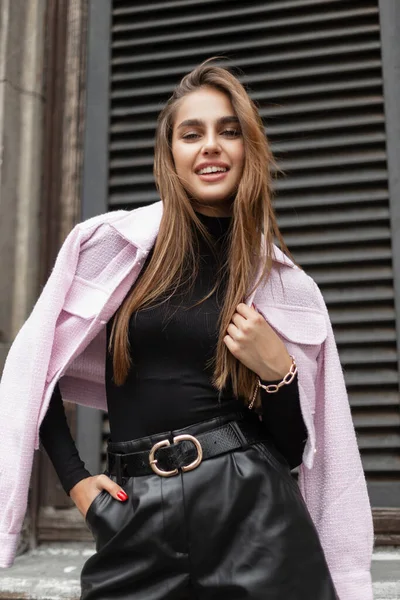 Happy beautiful fashionable woman urban model with a smile in black fashion casual elegant clothes with a pink jacket on shoulders near a vintage building