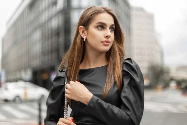Urban business female portrait of a beautiful stylish woman model in a fashion black dress with a trendy bag on the background of a modern city