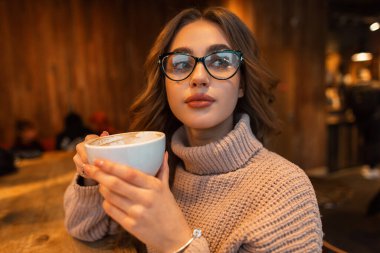 Indoors female portrait of a beautiful fashionable girl with trendy glasses in a stylish knitted vintage beige sweater holds a white mug and drinks coffee in a cafe