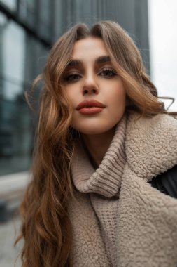 Urban female portrait of a beautiful stylish girl model with a clean face and curly hair in a fashion winter down jacket and a knitted vintage sweater in the city