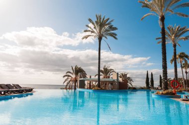 beautiful exotic view of a hotel with a pool, palm trees, sun beds and the ocean with a cloudy sky in Madeira island. Summer holidays in Funchal