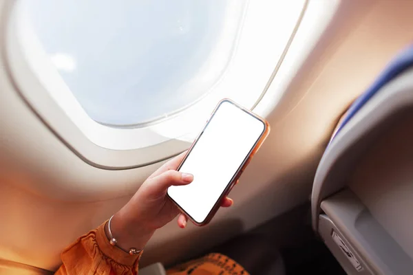 Professional business woman is flying in an airplane and using a smart phone near the porthole. Travel and smartphone mockup. Woman\'s hand holding a phone