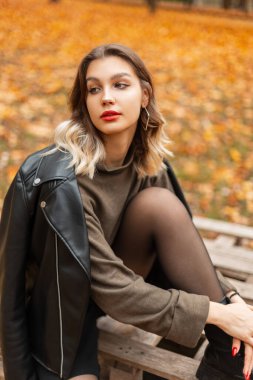 Moda yeşili kazaklı, siyah deri ceketli güzel bir kadın sonbahar parkında ahşap bir palette oturuyor.